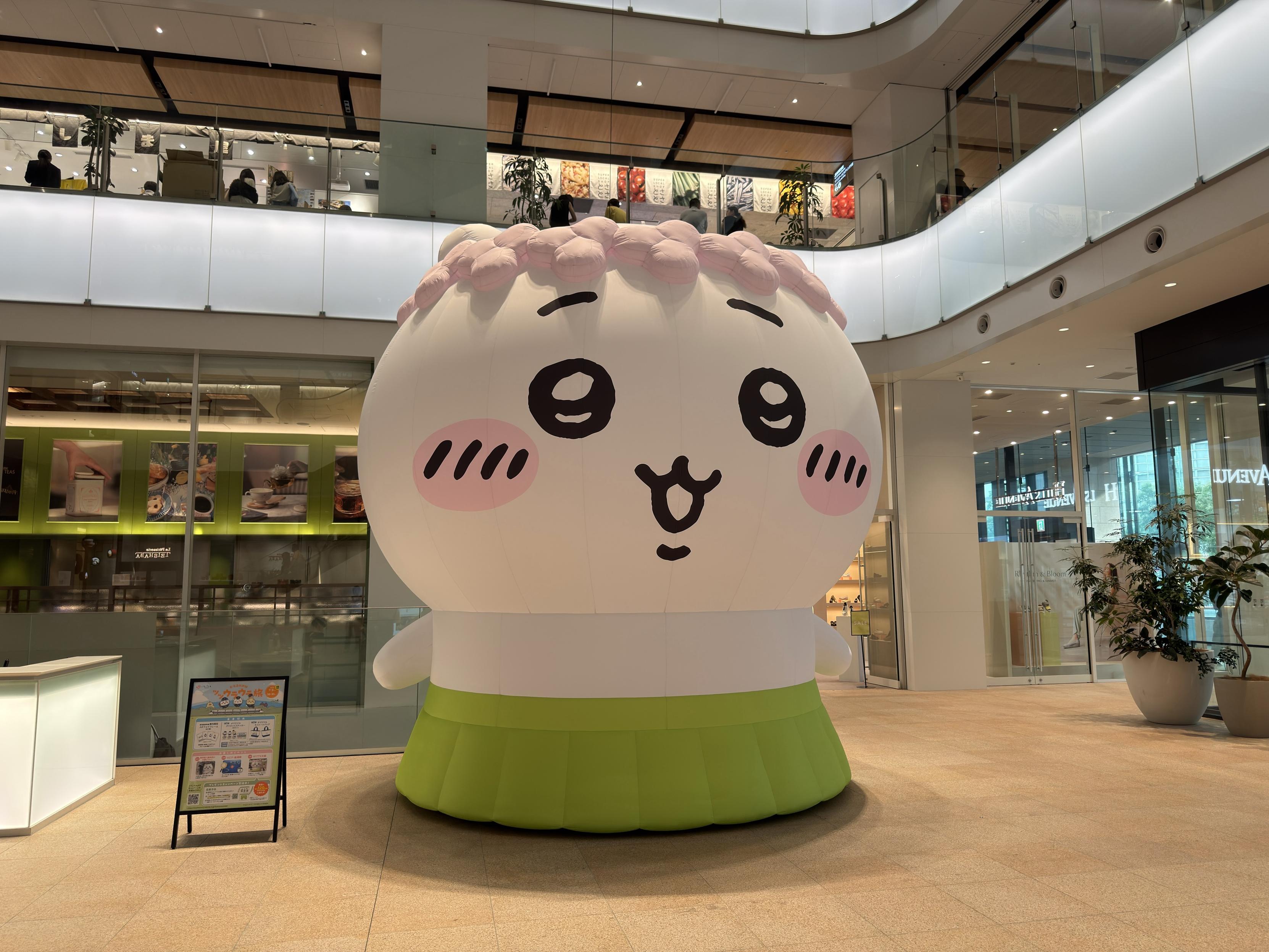A large, inflatable cartoon character resembling a cute rice ball with a smiling face, pink cheeks, and flower details on top, is prominently displayed indoors. The character is set against a modern backdrop with green accents and glass panels.